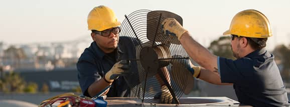 Two men wearing yellow hardhats and fixing a fan